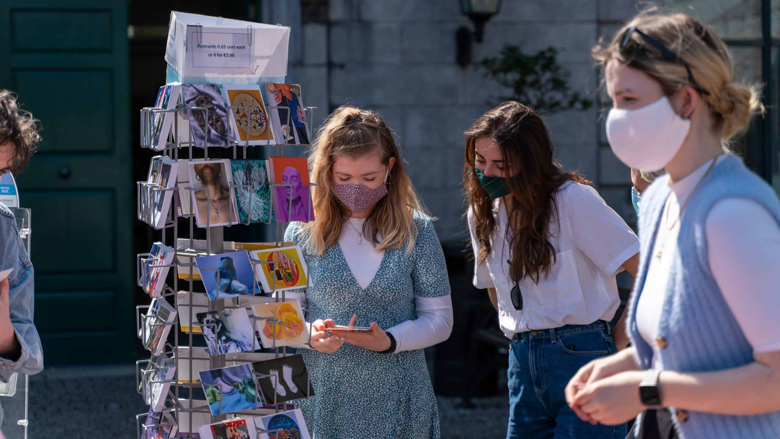 Two students look at a postcard beside a rack of postcards. Another student walks by in the foreground. Third Level Education